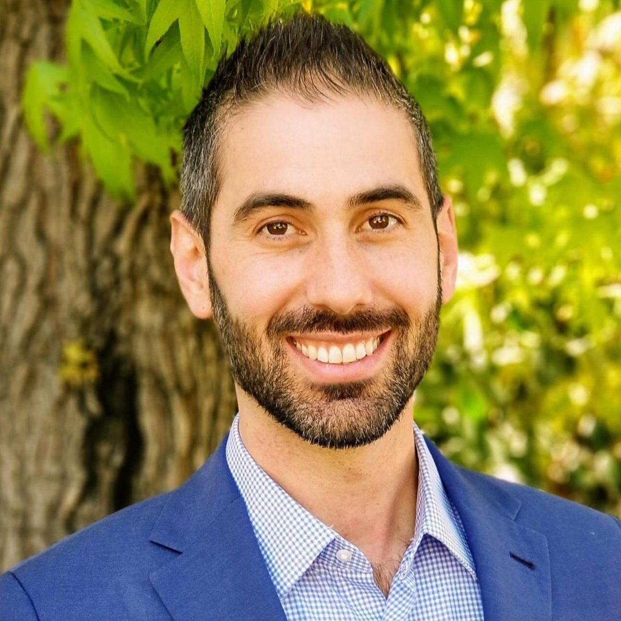 A man in a blue blazer stands outdoors with green leaves in the background.