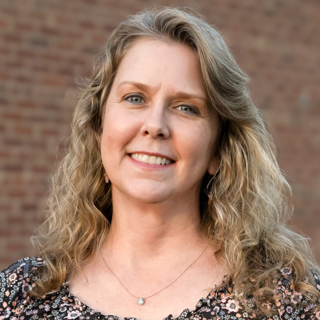 A woman with wavy blonde hair and a floral top stands smiling against a brick wall.