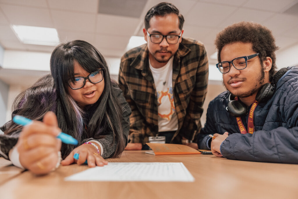 Three individuals are gathered around a table in an indoor setting, focused intently on a document in front of them.
