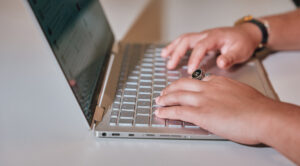 The image shows a close-up view of a laptop on a light-colored surface. A person's hands are poised over the keyboard, fingers positioned as if typing.