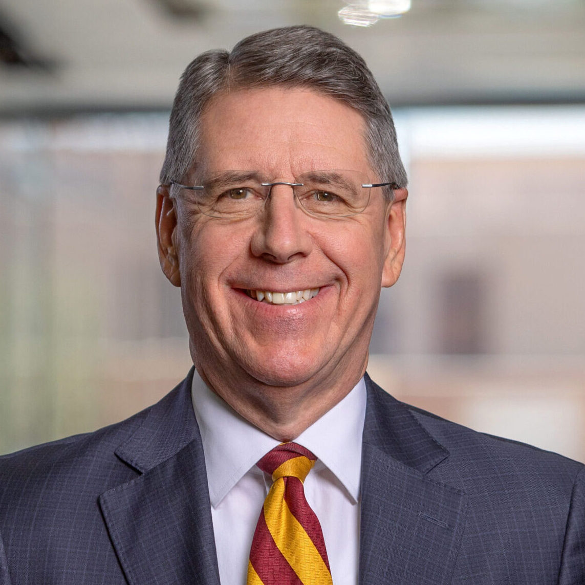 A man wearing a dark suit and a maroon and gold tie smiles at the camera.