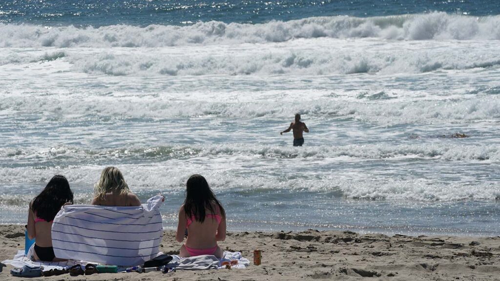 Three people sit on a beach and watch another person in the ocean.
