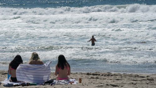 BeachandBay Three people sit on a beach and watch another person in the ocean.
